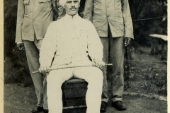 Zwei indische Soldaten stehen auf beiden Seiten eines Offiziers, der in einer weißen Uniform auf einem Stuhl sitzt