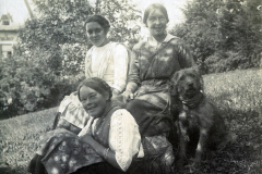 Drei Frauen in traditioneller Kleidung posieren mit Hunden im Freien in einer ländlichen Umgebung