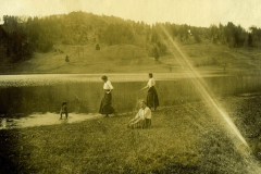 Darstellung einer ländlichen Szene mit mehreren Personen am Ufer eines ruhigen Gewässers in den frühen 1900er Jahren