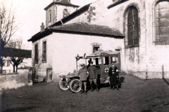 Ein historischer Moment mit einem Krankenwagen vor einer Kirche in ländlicher Umgebung