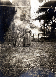 Frau im Garten steht mit einem Kind, umgeben von Bäumen und einem Gebäude, in einer historischen schwarz-weiß Aufnahme