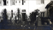 Eine Frau steht in einem Garten vor einem historischen Gebäude mit Fenstern und Pflanzen im Hintergrund