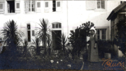 Eine Frau steht in einem Garten vor einem historischen Gebäude mit Fenstern und Pflanzen im Hintergrund