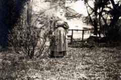 Frau im Garten steht mit einem Kind, umgeben von Bäumen und einem Gebäude, in einer historischen schwarz-weiß Aufnahme