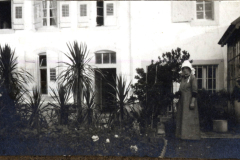 Eine Frau steht in einem Garten vor einem historischen Gebäude mit Fenstern und Pflanzen im Hintergrund