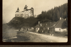 Burg auf einem Hügel inmitten einer bewaldeten Landschaft mit einer Straße und Gebäuden im Vordergrund