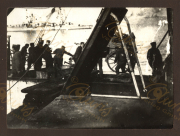 Soldaten in Uniform verladen schwere Ausrüstung auf ein Schiff. Die Szenerie spielt sich an einem Gewässer ab, möglicherweise während einer militärischen Operation im Ersten Weltkrieg. Die Männer wirken konzentriert und organisiert.
