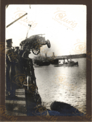 Marinepersonal beobachtet den Hublift eines Geschützes ins Wasser am Hafen. Die Szene zeigt das militärische Treiben während des Ersten Weltkriegs, mit Schiffen im Hintergrund und einer dramatischen Atmosphäre.