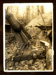 Das Bild zeigt ein Haubitzenrohr des Zuges 48 P.B. 11 in einem Feldlager, umgeben von Laub und einer gezelteten Struktur. Diese Aufnahme vermittelt einen Eindruck vom militärischen Alltag während des Ersten Weltkriegs in einem bewaldeten Gebiet.