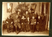 In einer festlich geschmückten Umgebung posiert eine Gruppe von Soldaten und Zivilisten um einen prächtigen Weihnachtsbaum. Die Männer in Uniform und die Frauen in eleganter Kleidung vermitteln eine Atmosphäre von Gemeinschaft und Feierlichkeit.
