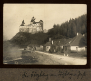 Burg auf einem Hügel inmitten einer bewaldeten Landschaft mit einer Straße und Gebäuden im Vordergrund