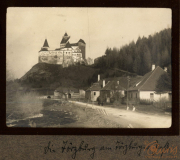 Burg auf einem Hügel inmitten einer bewaldeten Landschaft mit einer Straße und Gebäuden im Vordergrund