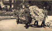 Soldaten tragen eine Kranzschleife in einer Gedenkzeremonie im Freien, umgeben von Blumen und Bäumen