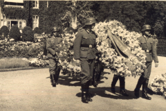 Soldaten tragen eine Kranzschleife in einer Gedenkzeremonie im Freien, umgeben von Blumen und Bäumen