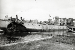 Ein stark beschädigtes Kriegsschiff liegt im Hafen, wobei mehrere Besatzungsmitglieder auf dem Deck stehen. Das Schiff weist schwere Zerstörungen am Heck und an den Aufbauten auf.