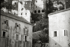 Die Aufnahme zeigt eine mediterrane Küstenstadt mit charakteristischer Steinarchitektur, einem markanten Kirchturm und Pferdewagen im Vordergrund. Die terrassenförmig an einem Hang gebauten Gebäude und der architektonische Stil deuten auf die Adria-Region hin.