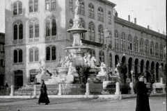 Ein großer Platz mit einem monumentalen Brunnen im Zentrum, umgeben von mehrstöckigen Gebäuden mit Arkadengängen im österreichisch-ungarischen Baustil. Zwei Personen in dunkler Kleidung sind auf dem gepflasterten Platz zu sehen.