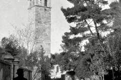 Die Aufnahme zeigt einen markanten Kirchturm mit spitzem Dach und Glockengeschoss in einer südeuropäischen Stadt. Auf der Straße sind mehrere Personen zu sehen, darunter eine uniformierte Gestalt im Vordergrund.