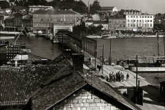 Blick über eine mediterrane Hafenstadt mit mehrstöckigen Gebäuden und bewaldeten Hügeln im Hintergrund. Im Hafen sind mehrere Kriegsschiffe und Marineeinrichtungen zu erkennen, während Marinepersonal auf den Kais versammelt ist.