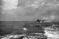 Ein deutsches Kriegsschiff der Kaiserlichen Marine fährt bei stürmischen Wetterbedingungen und hohem Seegang. Das Schiff ist in der Ferne zu erkennen, während im Vordergrund die aufgewühlte See mit schäumenden Wellenkämmen zu sehen ist.