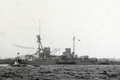 Ein großes Schlachtschiff der deutschen Kaiserlichen Marine liegt im Hafen, erkennbar an den charakteristischen Geschütztürmen und der militärischen Bauweise. Im Vordergrund ist ein kleineres Fahrzeug auf dem Wasser zu sehen.