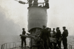 Marineangehörige in Uniform versammeln sich um ein Deckgeschütz auf einem deutschen Kriegsschiff. Die Aufnahme zeigt den Schornstein und die Takelage des Schiffes bei ruhiger See.
