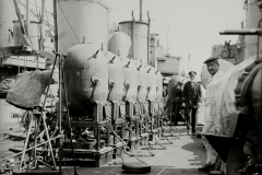 Mehrere Seeminen sind auf dem Deck eines Kriegsschiffes aufgereiht, während Marineoffiziere in Uniformen daneben stehen. Die zylindrischen Minen mit ihren charakteristischen Kontakthörnern sind zur Verladung oder Installation bereit.