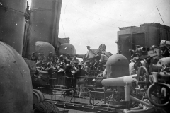 Marineangehörige der Kaiserlichen Marine musizieren mit Blasinstrumenten auf dem Deck eines Kriegsschiffes. Die Aufnahme zeigt Geschütze, Aufbauten und technische Ausrüstung des Schiffes im Hintergrund.