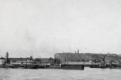 Die Aufnahme zeigt eine befestigte Hafenanlage mit Molen, Kränen und militärischen Einrichtungen vor einer Festung auf einem Hügel. Im Vordergrund sind Hafenbecken und Pier-Anlagen der Kaiserlichen Marine zu erkennen.