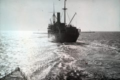 Ein deutsches Kriegsschiff mit charakteristischem Schornstein und Masten fährt auf ruhiger See. Die Aufnahme wurde vom Deck des U-Bootes S.M. U 39 aus gemacht, erkennbar am Reling im Vordergrund.