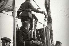Mehrere Marineoffiziere und Besatzungsmitglieder der Kaiserlichen Marine auf der Brücke eines Kriegsschiffs. Ein Offizier mit Mütze steht am Mast und bedient nautische Ausrüstung, während andere Besatzungsmitglieder in Uniformen verschiedene Positionen auf der Brücke einnehmen.