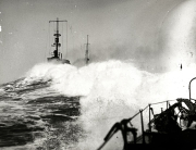 Ein Kriegsschiff kämpft sich durch schwere See, während hohe Wellen über Bug und Aufbauten brechen. Die Aufnahme wurde von einem anderen Schiff aus gemacht, dessen Deck und Ausrüstung im Vordergrund zu sehen sind.