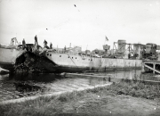 Ein stark beschädigtes Kriegsschiff liegt im Hafen, wobei mehrere Besatzungsmitglieder auf dem Deck stehen. Das Schiff weist schwere Zerstörungen am Heck und an den Aufbauten auf.
