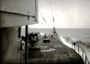Aufnahme vom Deck eines deutschen U-Bootes während der Fahrt mit Blick auf das Deckgeschütz und die Periskopanlage. Das U-Boot durchpflügt bei ruhiger See das Wasser, wobei Bug und Aufbauten deutlich sichtbar sind.