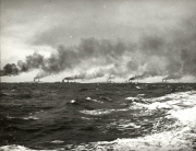 Mehrere deutsche Kriegsschiffe der Kaiserlichen Marine fahren in Formation am Horizont und erzeugen charakteristische Rauchsäulen. Die Aufnahme zeigt schweren Seegang mit schaumgekrönten Wellen im Vordergrund.