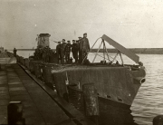 Ein deutsches U-Boot liegt im Hafen, während die Besatzung in Uniformen auf dem Deck und Turm versammelt ist. Das Foto zeigt das Fahrzeug von der Seite mit deutlich sichtbarem Kommandoturm und Periskop.