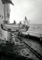 Ein Matrose hält eine kleine Figur oder ein Maskottchen auf dem Deck eines U-Boots. Im Hintergrund sind weitere Kriegsschiffe auf See zu erkennen.