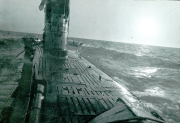 Das Deck eines deutschen U-Bootes bei rauer See mit deutlich sichtbarem Turm und starkem Wellengang. Die Aufnahme zeigt das nasse Holzdeck und die schwierigen Seebedingungen während der Fahrt.
