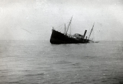 Ein Dampfschiff mit deutlicher Schlagseite nach Steuerbord liegt sinkend im Wasser. Das Schiff zeigt Masten, einen Schornstein und Aufbauten eines typischen Handelsschiffes der Zeit des Ersten Weltkriegs.