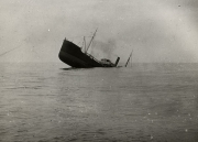 Ein Handelsschiff mit Dampfschornstein und Segeltakelung sinkt mit starker Schlagseite nach Backbord. Das Schiff ist bereits tief im Wasser und die Masten ragen schräg aus dem Meer.