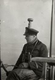 Ein Offizier der Kaiserlichen Marine in Uniform und Schirmmütze steht mit einem Fernglas auf der Brücke eines U-Bootes. Im Hintergrund ist das Sehrohr des Unterseebootes erkennbar.