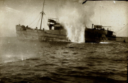 Ein Dampfschiff wird durch einen Torpedotreffer versenkt, eine große Wassersäule steigt zwischen Bug und Heck auf. Das Schiff ist bereits stark beschädigt und beginnt zu sinken.