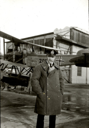 Ein Marineoffizier in Uniform steht vor einem Seeflugzeug mit der Kennung 207 und dem Balkenkreuz. Im Hintergrund sind Werftgebäude und weitere Flugzeuge zu erkennen.