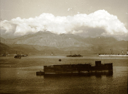 Ein U-Boot liegt im Vordergrund einer Bucht, während mehrere Kriegsschiffe und kleinere Fahrzeuge im Hintergrund vor einer dramatischen Berglandschaft ankern. Die Aufnahme zeigt den Marinestützpunkt Cattaro oder Pola während des Ersten Weltkriegs.