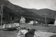 Blick auf den Hafen von Cattaro mit einer orthodoxen Kirche im Hintergrund und der charakteristischen Berglandschaft der Bucht von Kotor. Die Aufnahme zeigt Hafeninfrastruktur mit Kaimauern und Masten im Vordergrund.