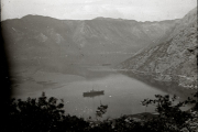 Panoramablick auf die fjordartige Bucht von Cattaro mit einem Kriegsschiff in der Mitte der ruhigen Gewässer. Die charakteristische Berglandschaft der montenegrinischen Küste umrahmt die strategisch wichtige Marinebasis der k.u.k. Marine.