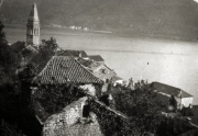 Die Aufnahme zeigt eine mediterrane Küstenlandschaft mit charakteristischen Ziegeldächern und einem markanten Kirchturm. Im Hintergrund erstreckt sich eine weite Meeresbucht mit Bergen am Horizont.