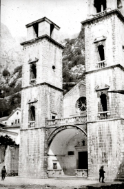 Die romanische Kathedrale des Heiligen Tryphon in Kotor zeigt ihre markante Doppelturmfassade aus hellem Steinmauerwerk. Im Vordergrund sind Personen zu erkennen, die den Maßstab des monumentalen Kirchenbaus verdeutlichen.