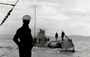 Ein deutsches U-Boot der Kaiserlichen Marine fährt aufgetaucht auf See, während mehrere Besatzungsmitglieder auf dem Turm und Deck stehen. Im Vordergrund ist die Silhouette eines Marinesoldaten mit Schirmmütze zu sehen, der das Geschehen vom Deck eines anderen Schiffes aus beobachtet.