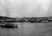 Die Aufnahme zeigt einen Hafen mit einem prächtigen mehrstöckigen Gebäude im Hintergrund und verschiedenen Kriegsschiffen im Hafenbecken. Links im Vordergrund sind kleinere Hafenfahrzeuge und Pontons zu erkennen.